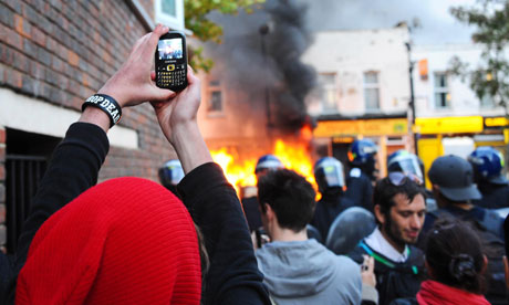 Foto van iemand die met een telefoon een brand in een straat fotografeert, met omstanders en agenten met helmen op de achtergrond.