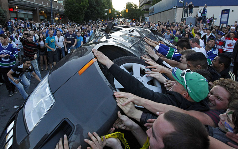 Een groep jonge mannen in Canucks-shirts duwt een auto op zijn kant, omringd door een grote menigte toeschouwers.
