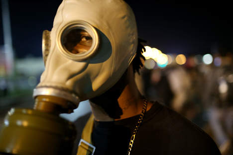Een demonstrant met een ouderwets gasmasker op staat ’s avonds op straat, met wazige lichten op de achtergrond.