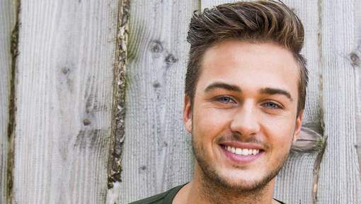 Portret van een lachende jonge man met opgeschoren donkerblond haar voor een houten schutting, vermoedelijk Dave Roelvink over wie de tekst ernaast gaat.