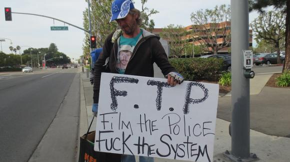 Een man met een pet staat langs een straat met een kartonnen bord met de tekst “F.T.P. Film the police, fuck the system”.