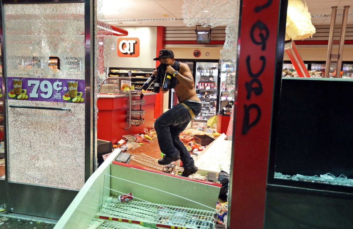 Een man met bedekt gezicht en flessen in zijn handen stapt door de ingeslagen ruit van een geplunderde QuikTrip-winkel. Op de vloer liggen omgegooide schappen en koopwaar.