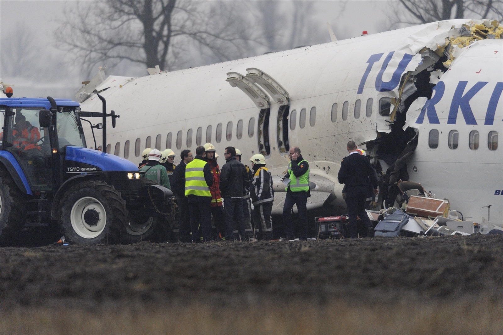 Wrak van vlucht TK1951 in een weiland: de romp met de tekst Turkish is opengescheurd en ervoor staan brandweerlieden, hulpverleners in hesjes en een tractor.