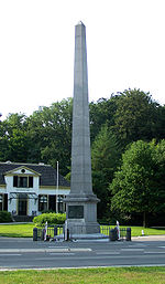 Het monument De Naald aan de Loolaan in Apeldoorn, een hoge stenen obelisk op een grasveld met bomen en een wit huis op de achtergrond.