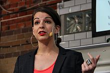 Anita Sarkeesian geeft een lezing met een headsetmicrofoon, voor een bakstenen muur.