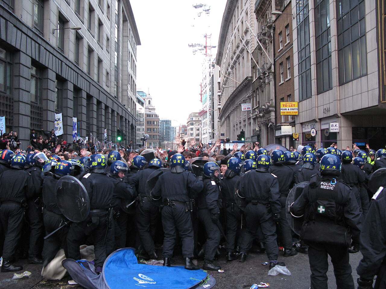 Foto van een rij politieagenten met helmen en schilden die in een Londense straat een groep demonstranten insluit, van wie velen hun handen opsteken; volgens de bestandsnaam bij het G20-klimaatkamp.