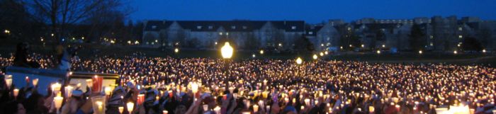 Een grote menigte met brandende kaarsen op een grasveld bij avond tijdens de wake na het schietincident op Virginia Tech, met universiteitsgebouwen op de achtergrond.