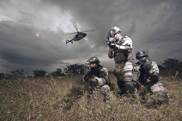 Zwaar bewapende soldaten van de Mexicaanse anti-drugseenheid in camouflagekleding en met helmen zitten gehurkt in hoog gras, terwijl er een helikopter onder een donkere lucht overvliegt.