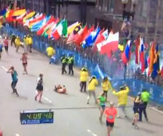 Stilstaand beeld uit een video van de finish van de Boston Marathon vlak na de explosie: lopers en vrijwilligers in gele hesjes bij een rij vlaggen, een loper op de grond en een wedstrijdklok.