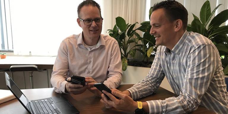 Twee lachende mannen in overhemd zitten aan een tafel met een laptop en kijken samen op hun smartphones.