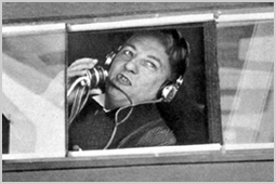 Zwart-witfoto van Willem Vogt met koptelefoon en microfoon, die door het raam van een KLM-vliegtuig verslag doet.