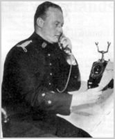 Zwart-witfoto van hoofdinspecteur G.D.J. Vrijdag uit Hilversum in uniform, die aan een bureau zit te telefoneren en een papier in zijn hand houdt.