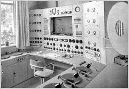 Zwart-witfoto van de ANRO-studio: een regieruimte met een grote wand vol knoppen, meters en schakelaars, een bureaustoel en draaitafels.