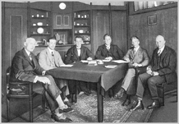Zwart-witfoto van het comité van de Hilversumsche Draadlooze Omroep in 1925: zes mannen in pak rond een tafel in een kamer met boekenkasten.