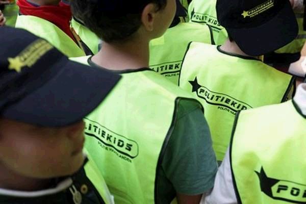 Close-up van een groep kinderen met donkere petten en fluorescerende gele hesjes met het opschrift “Politiekids”, van achteren gezien.