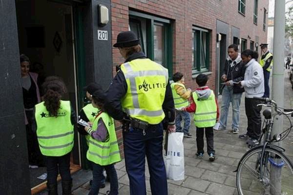 Kinderen in gele Politiekids-hesjes staan met een politieagent bij de voordeur van een rijtjeshuis in een stadswijk, terwijl verderop andere kinderen en volwassenen op de stoep staan.
