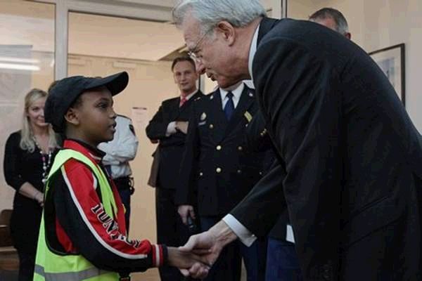 Een oudere man in pak schudt een jongen met pet en geel hesje de hand, terwijl op de achtergrond politiemensen in uniform en andere aanwezigen toekijken.