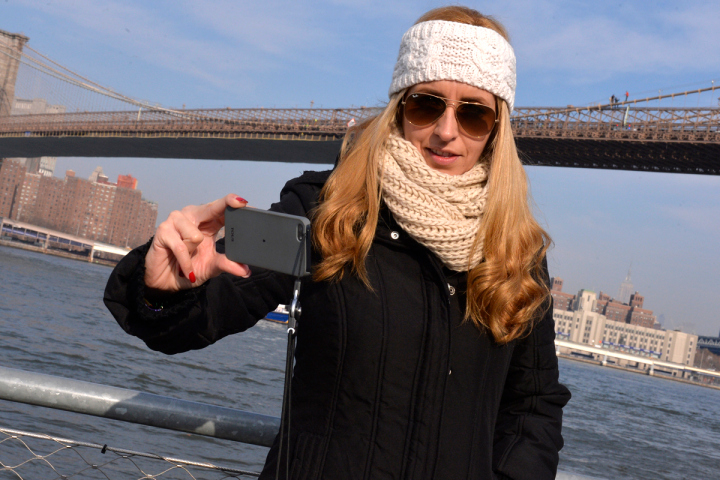 Een blonde vrouw met zonnebril en witte hoofdband maakt een selfie met haar telefoon aan het water, met de Brooklyn Bridge op de achtergrond.