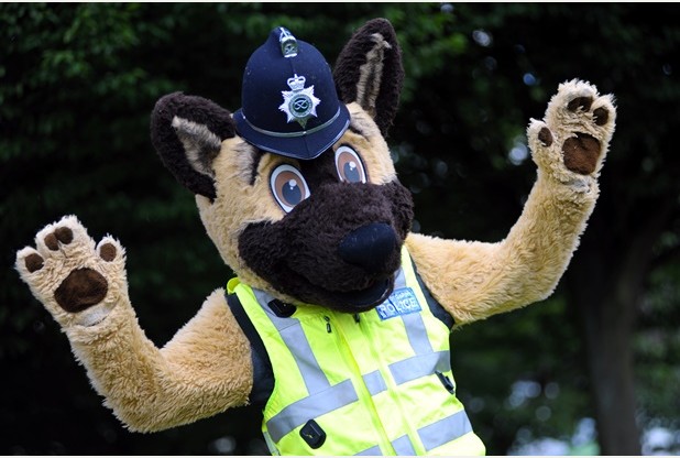 Politiehondmascotte in een pluche pak met politiehelm en geel hesje, met beide poten omhoog.