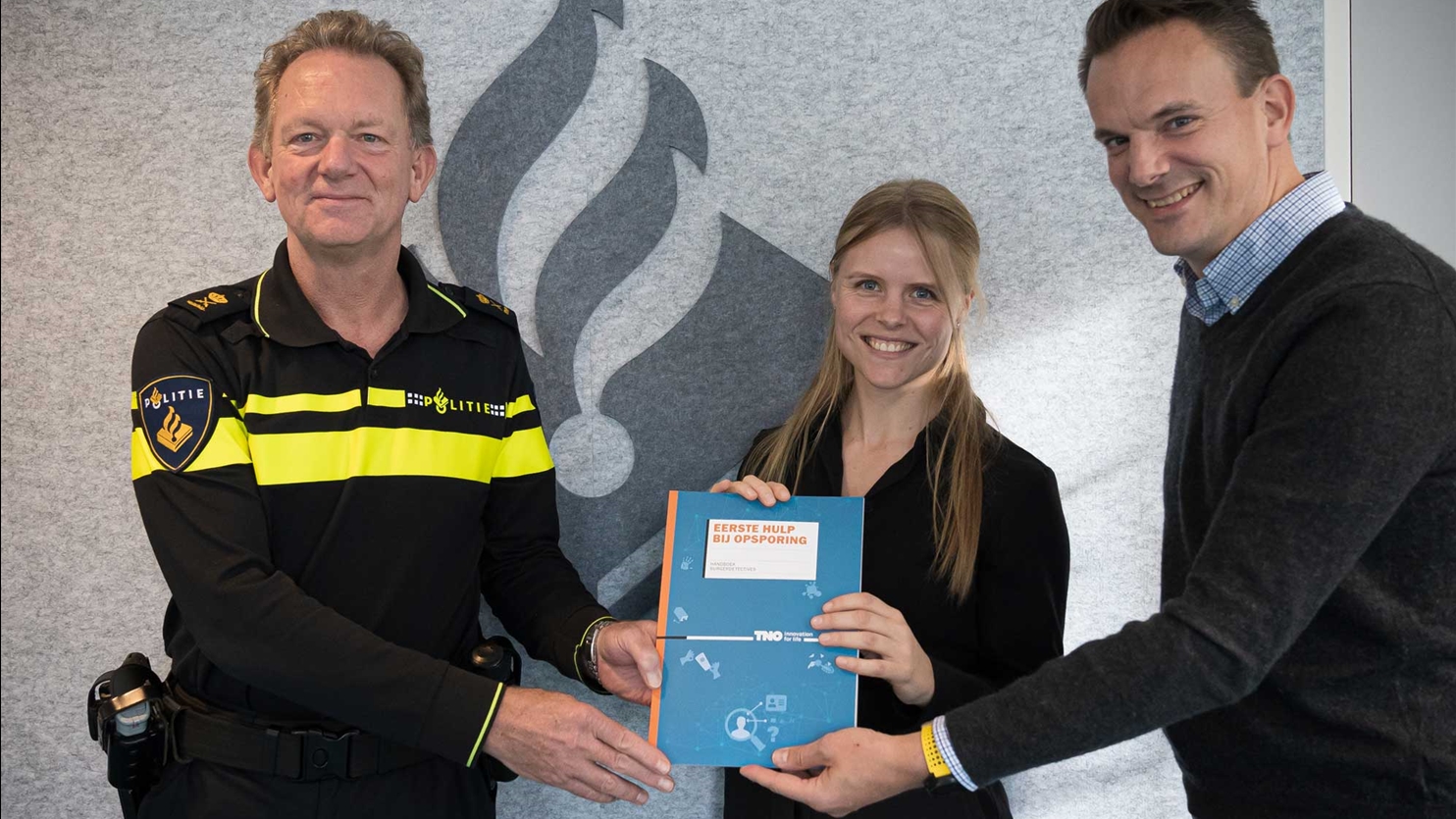 Overhandiging van het handboek “Eerste Hulp bij Opsporing”: van links naar rechts Fred Westerbeke (politie), Shanna Wemmers en Arnout de Vries (TNO) met het boek tussen hen in.